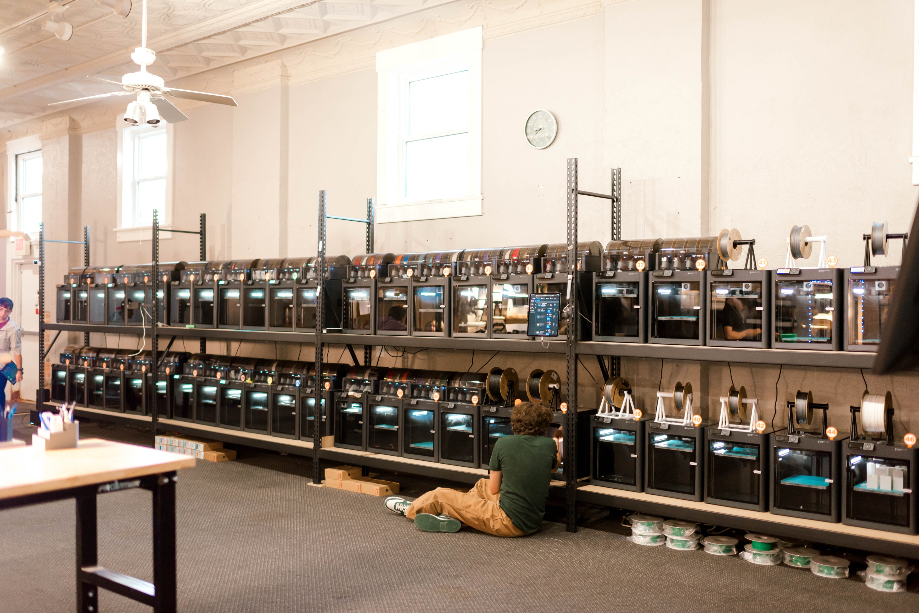 Long row of electronic 3d printer devices on shelves in a room with a person sitting on the floor.